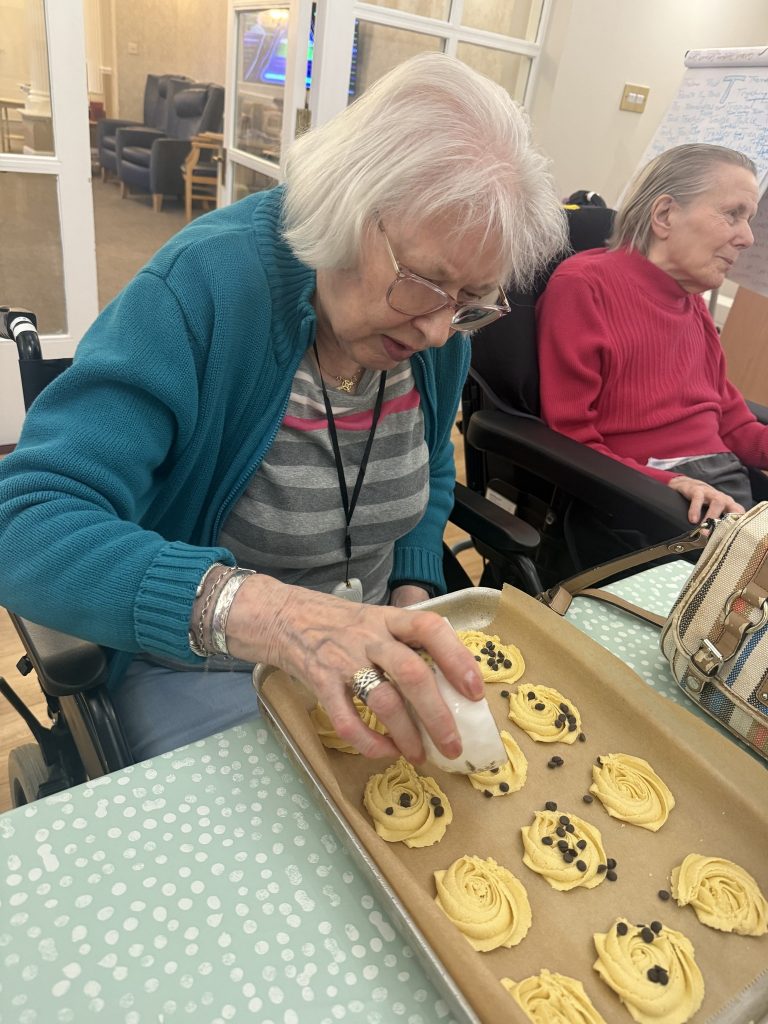 Elderly woman sprinkling chocolate chips onto unbaked cookies at a table.