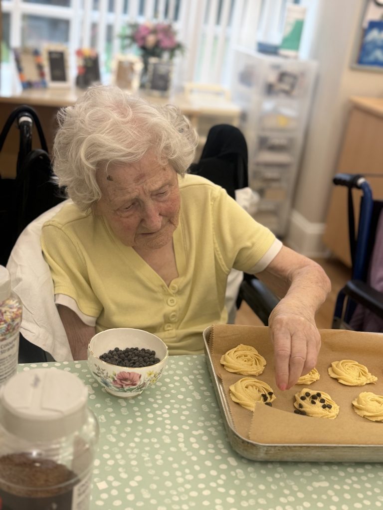 Elderly woman placing chocolate chips onto cookies beside a bowl of ingredients.