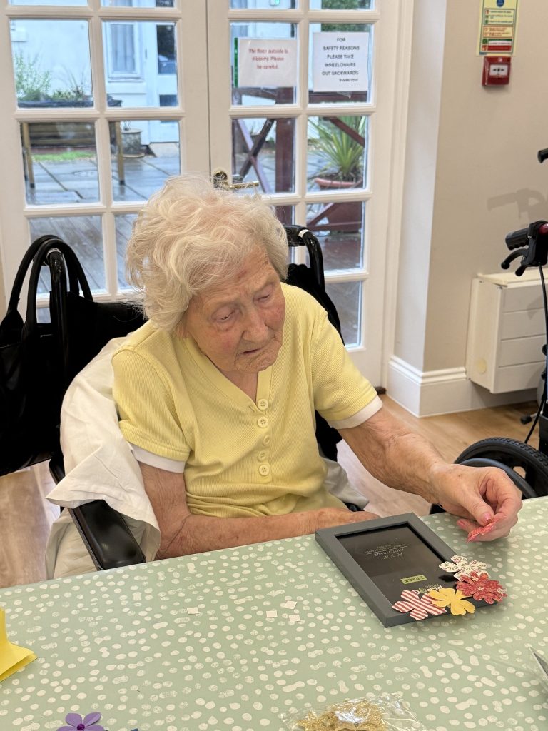 Elderly woman adding flower decorations to a photo frame at a craft table.