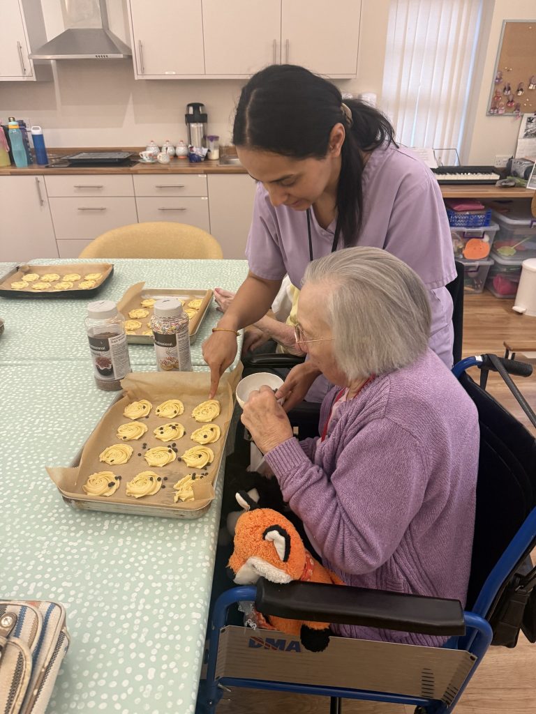Caregiver assisting an elderly woman in decorating cookies on a baking tray.
