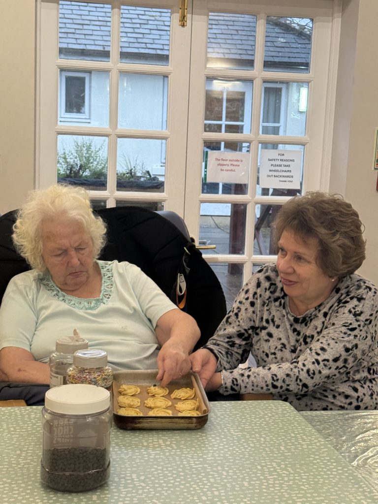Two elderly women decorating cookies together at a table with trays and toppings.