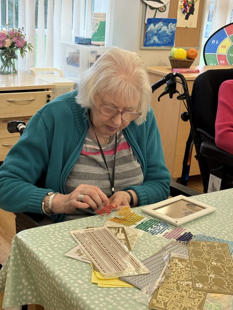 Elderly woman placing colorful gem stickers on a craft project at a table.