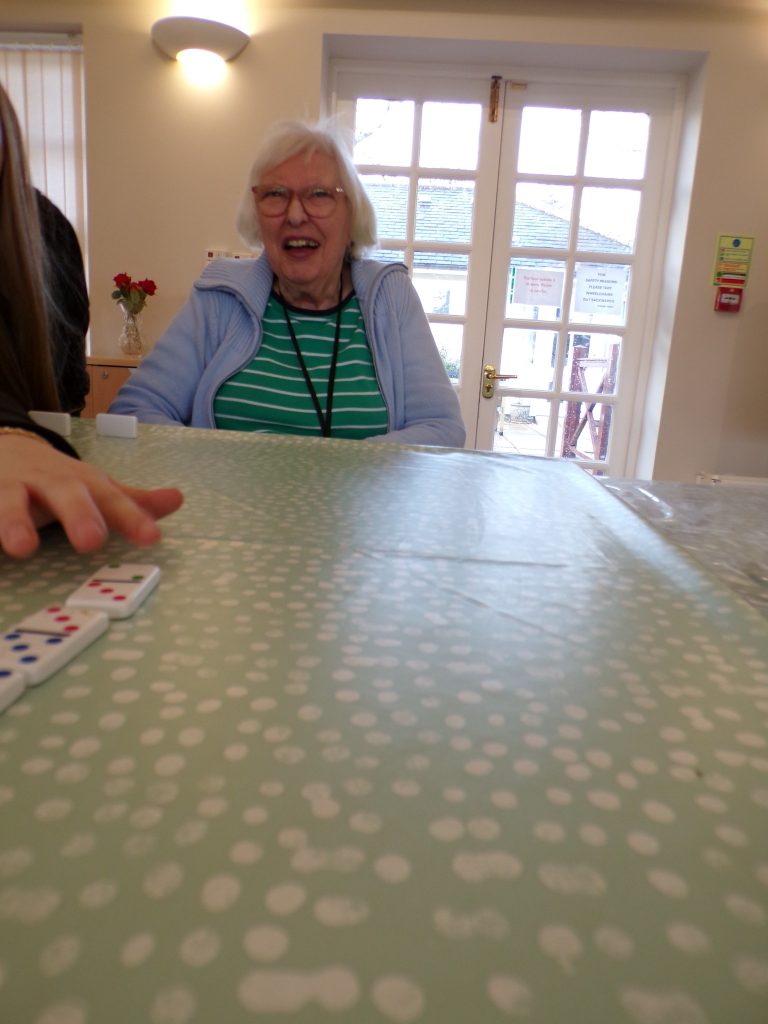 Elderly woman smiling while playing dominoes at a table.