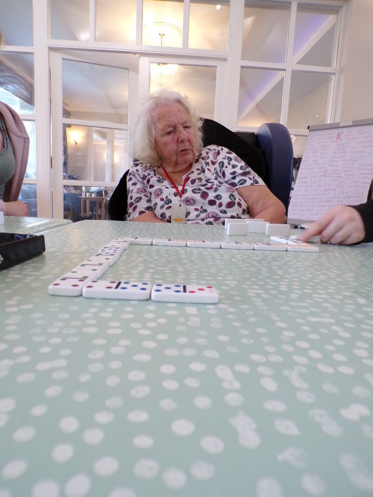 Elderly woman playing dominoes at a table with others assisting.