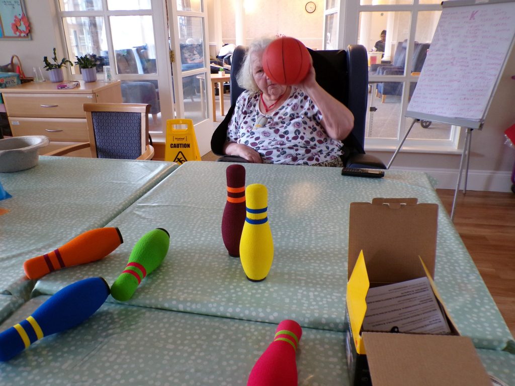 Elderly woman tossing a soft ball at colorful bowling pins on a table.