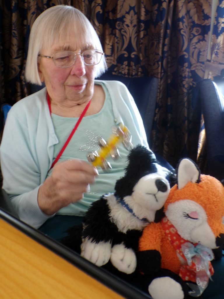 Elderly woman playing with a bell toy beside plush animals.