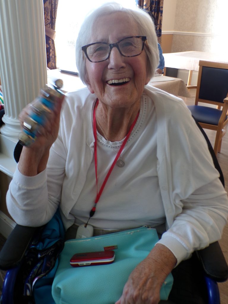 Smiling elderly woman shaking a small percussion instrument.
