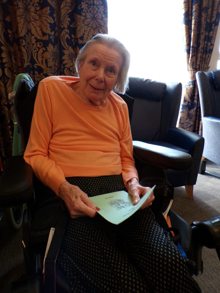 Elderly woman seated, holding a booklet and smiling.