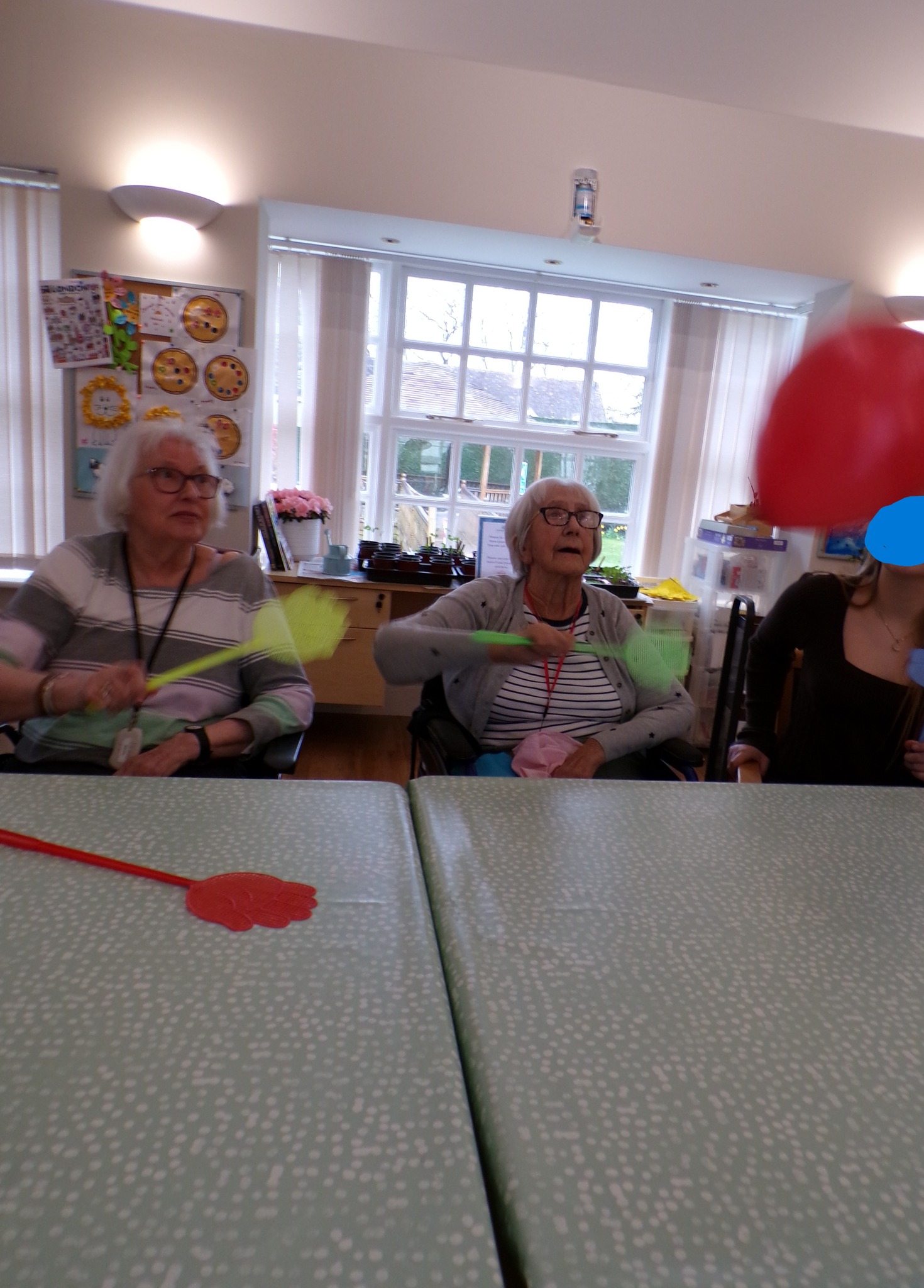 Elderly participants at a table batting a red balloon with paddles during a group activity.