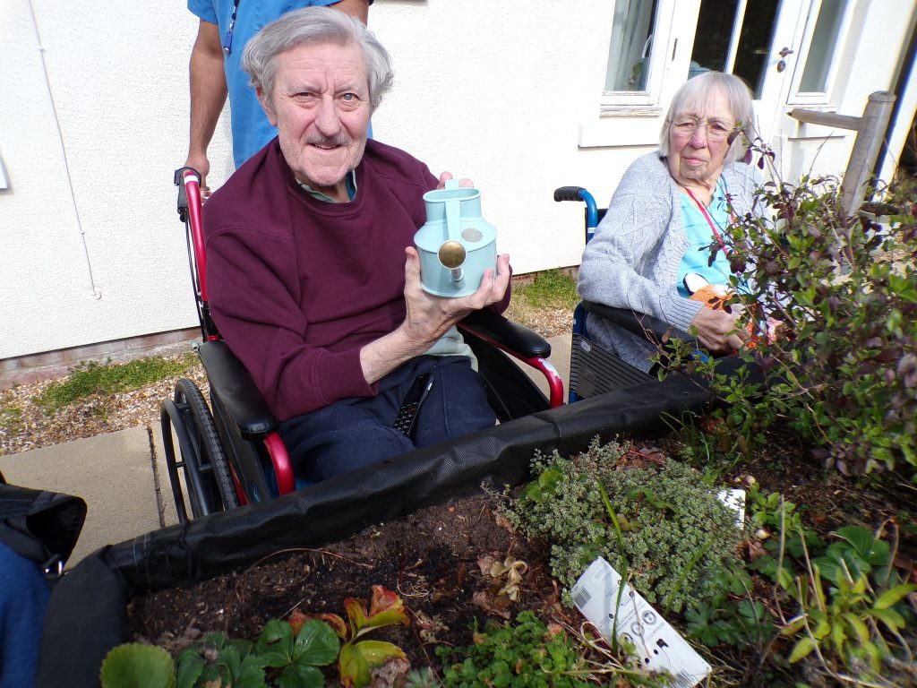 Elderly man in wheelchair holding a watering can next to a raised garden bed, with another woman nearby.