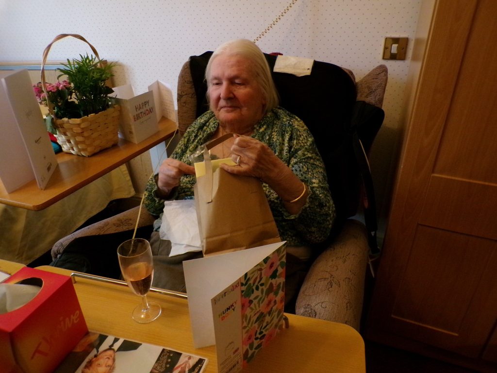 Elderly woman sitting in a chair opening a gift bag beside birthday cards and a drink.