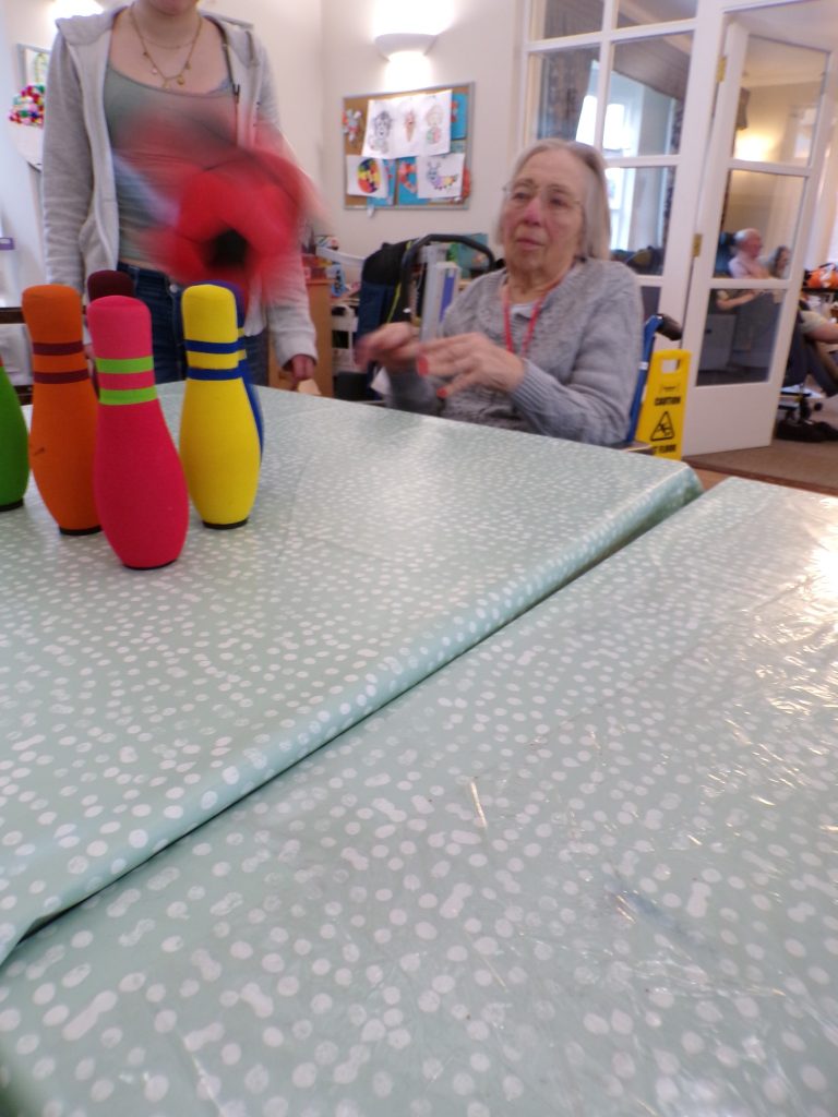 Older woman in a wheelchair reaching toward colorful bowling pins during a tabletop game.