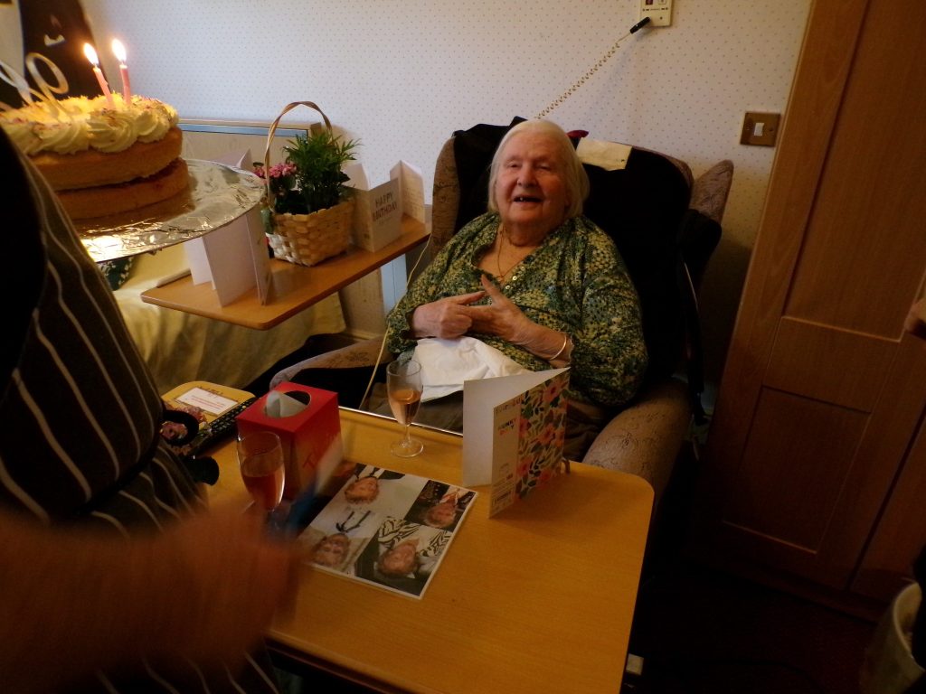 Elderly woman smiling in a chair as a birthday cake with candles is presented, with cards and a drink on the table.