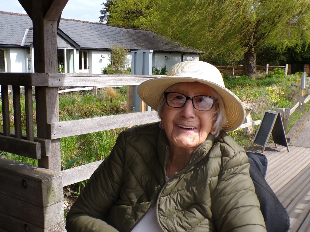Elderly woman wearing a hat and glasses, smiling outdoors in a garden setting.