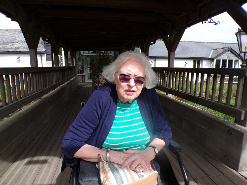 Elderly woman in a wheelchair wearing sunglasses, sitting on a wooden outdoor walkway.