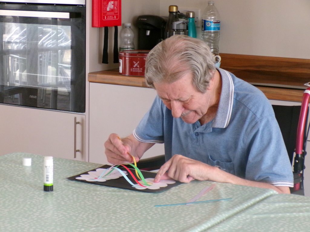 Elderly man concentrating while doing a colorful paper craft at a table.