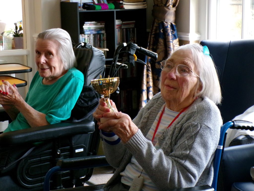 Elderly woman in a wheelchair holding a small trophy, smiling, with another woman beside her.