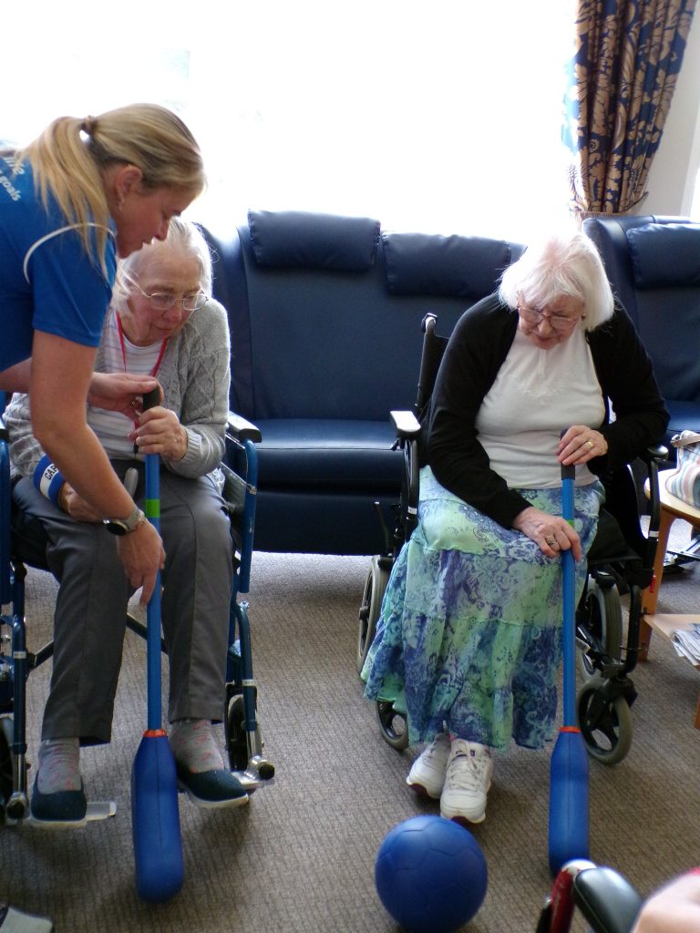 Two elderly women in wheelchairs playing a seated bowling game with a caregiver assisting.