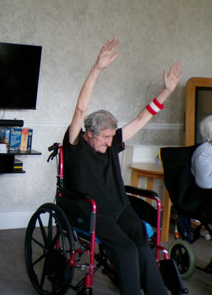 Elderly man in a wheelchair raising both arms during an activity session.