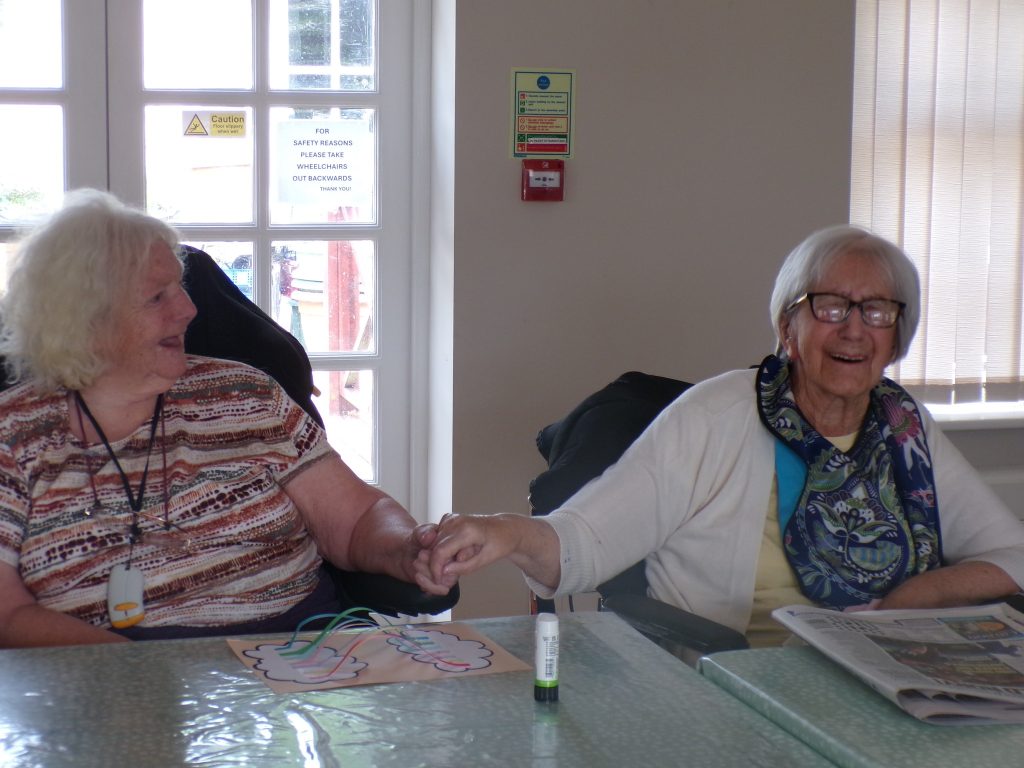 Two elderly women sitting at a table, holding hands and smiling during an activity.