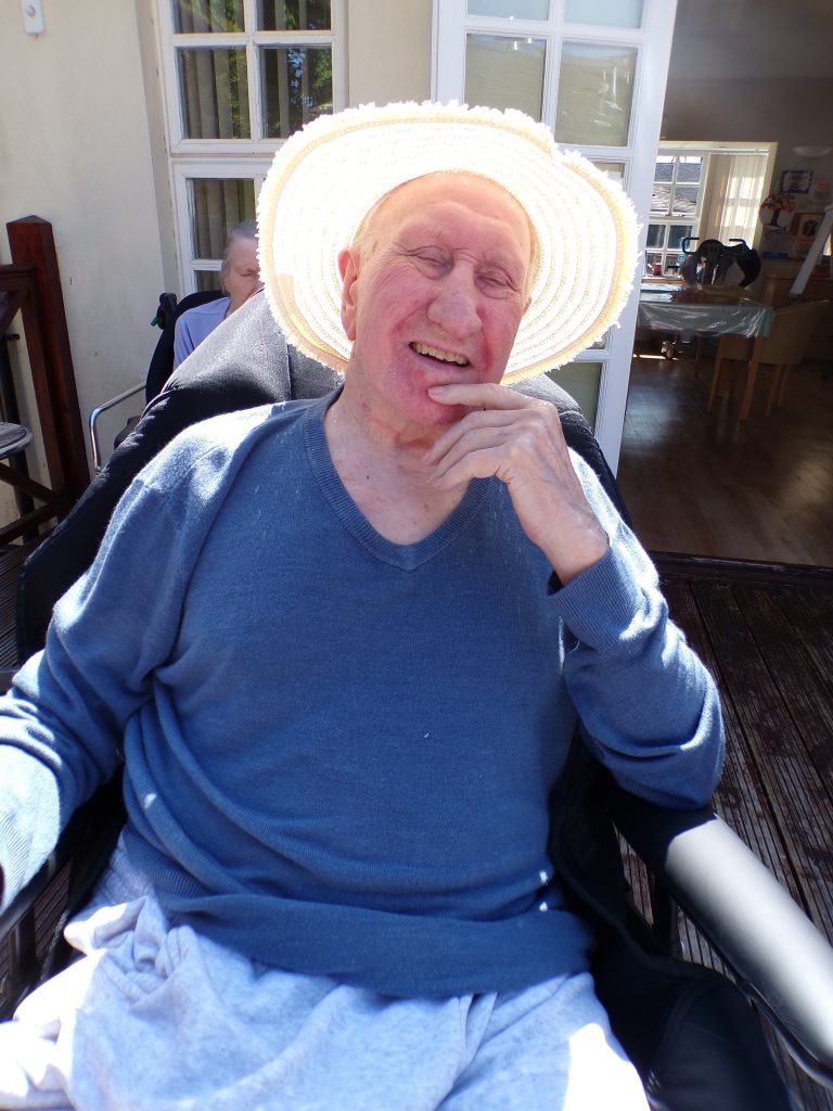 Elderly man in a sun hat smiling while sitting outside near a doorway.