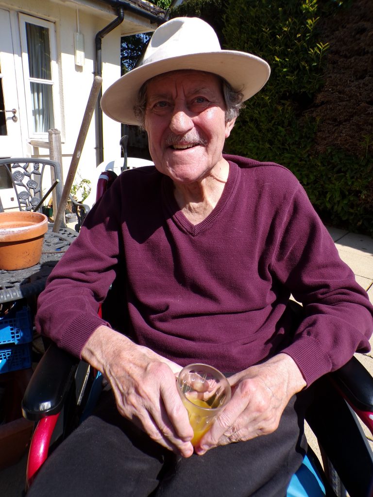 Elderly man in a hat sitting outdoors, smiling and holding a drink.