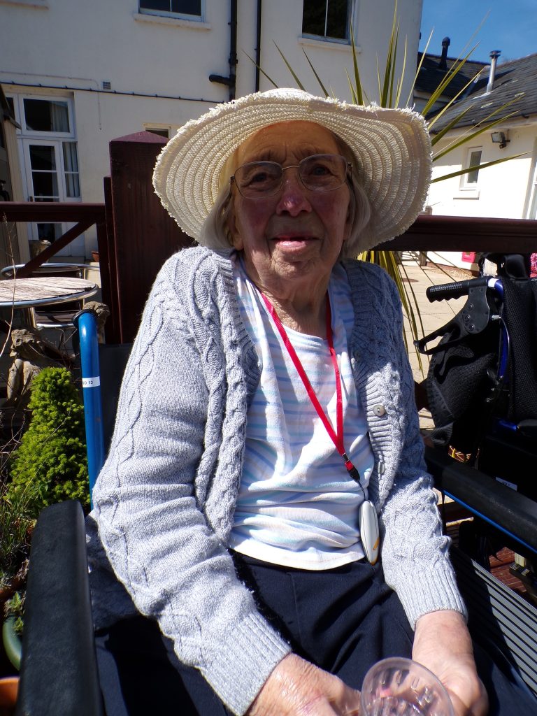 Elderly woman wearing glasses and a wide-brimmed hat sitting outside with a drink.