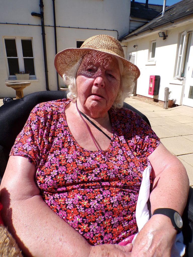 Elderly woman in a floral dress and straw hat sitting outdoors in bright sunlight.