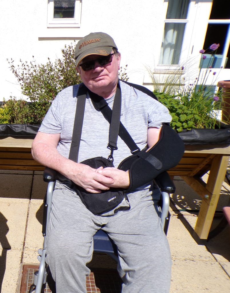 Elderly man wearing a cap and arm sling sitting outside on a sunny day.