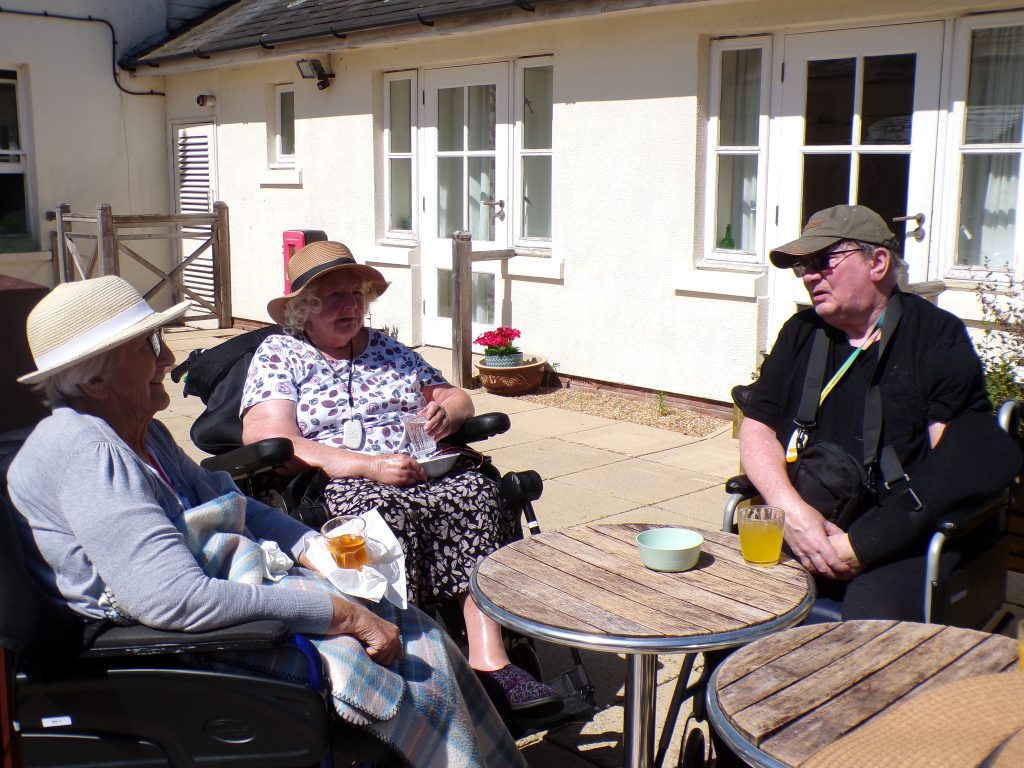 Three elderly people sitting outside in the sun, chatting around a small table with drinks.