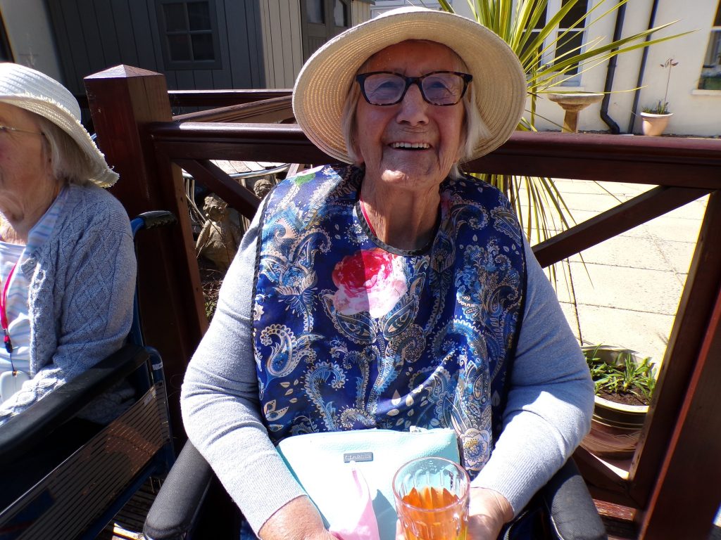 Elderly woman wearing glasses and a hat, smiling while holding a drink outdoors.