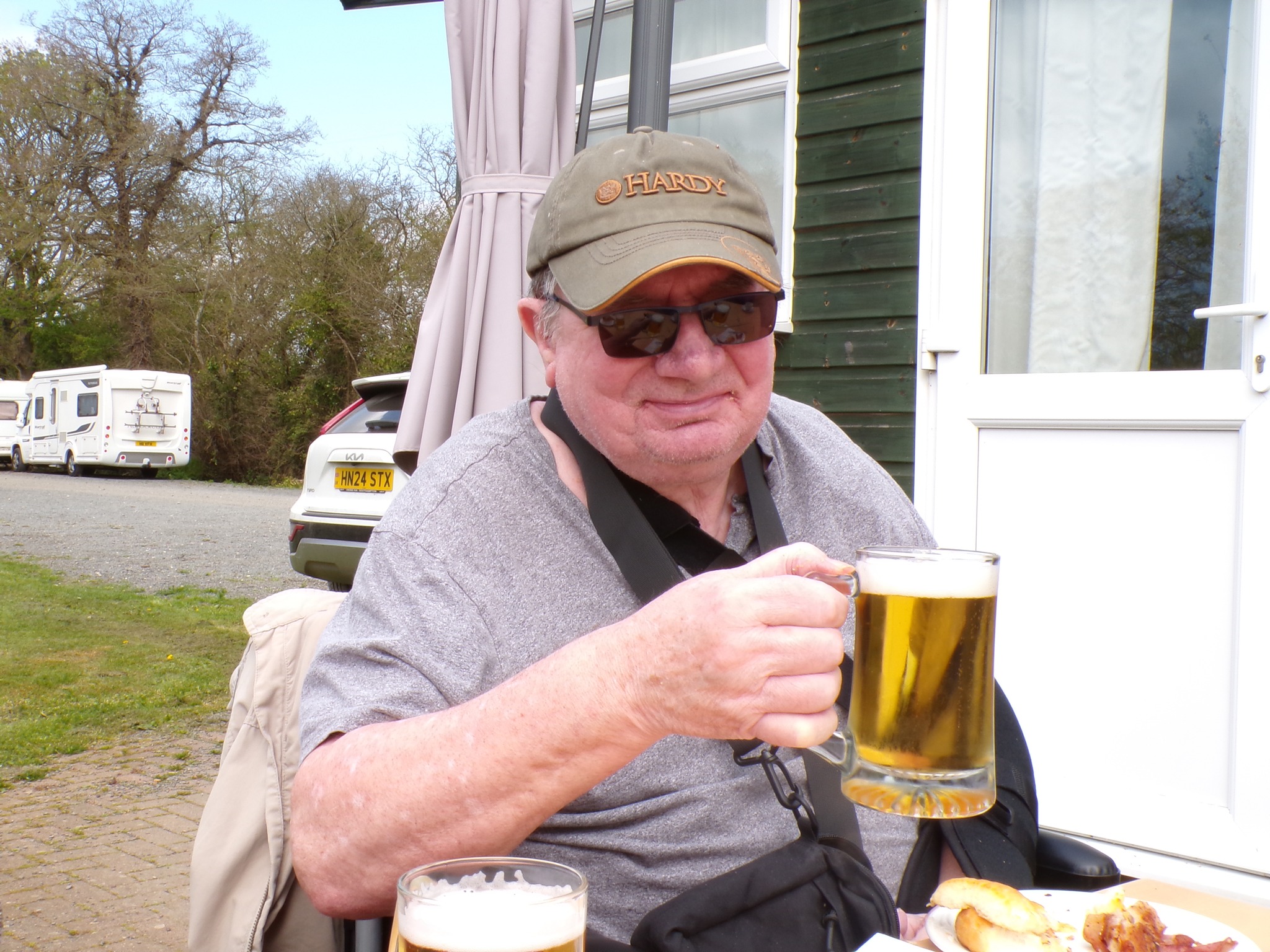 Man sitting outdoors holding a glass of beer, with a parked vehicle in the background.