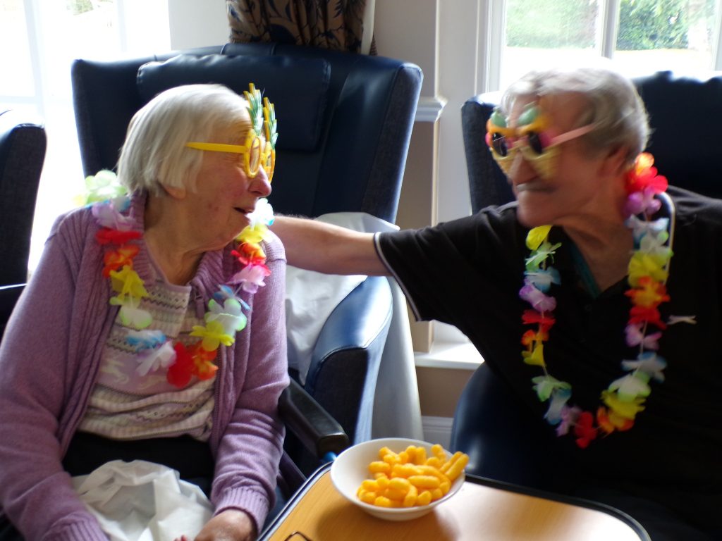 Two elderly people wearing fun sunglasses and flower leis indoors.