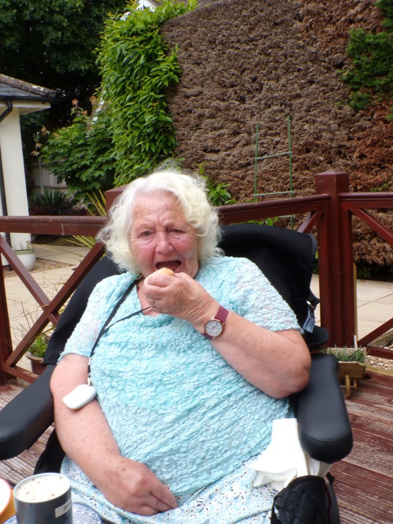 Elderly woman sitting outdoors in a wheelchair eating a snack.