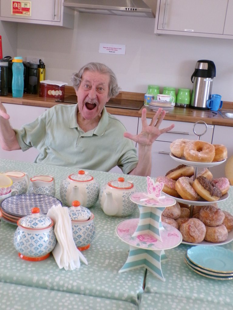 Elderly man smiling excitedly beside donuts and tea sets.