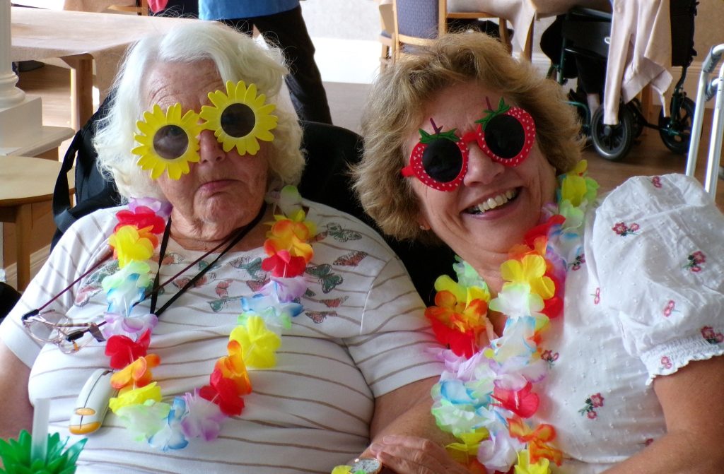 Two women smiling together wearing novelty sunglasses and leis.