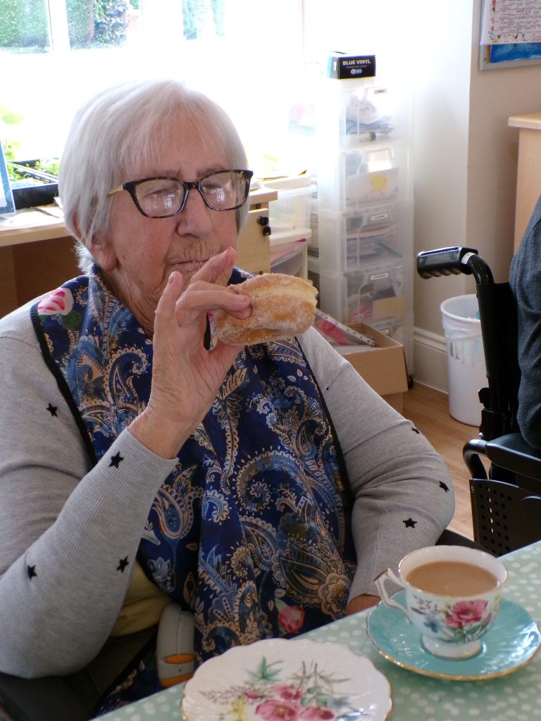 Elderly woman enjoying tea and a sugared donut at a table.