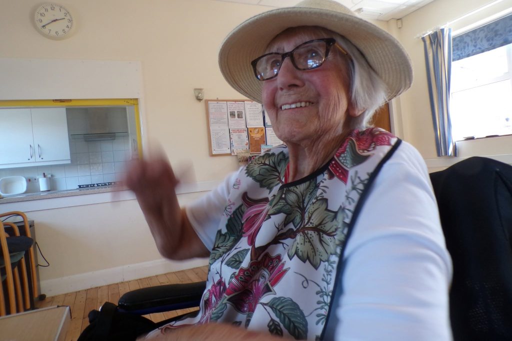 Smiling elderly resident wearing a sun hat and glasses, enjoying time in a bright nursing home lounge