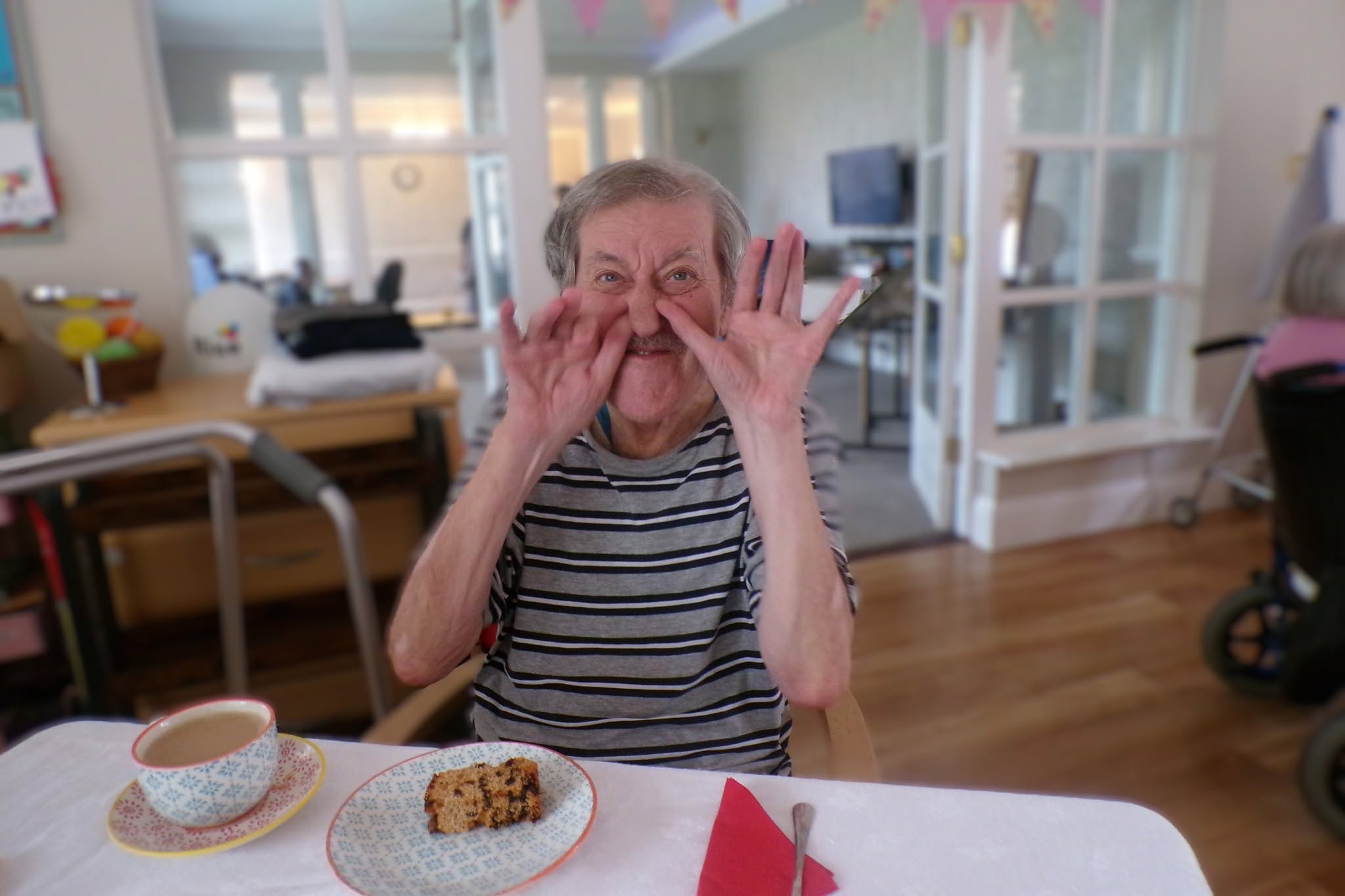 Cheerful elderly resident making a playful gesture while enjoying refreshments in the care home lounge.